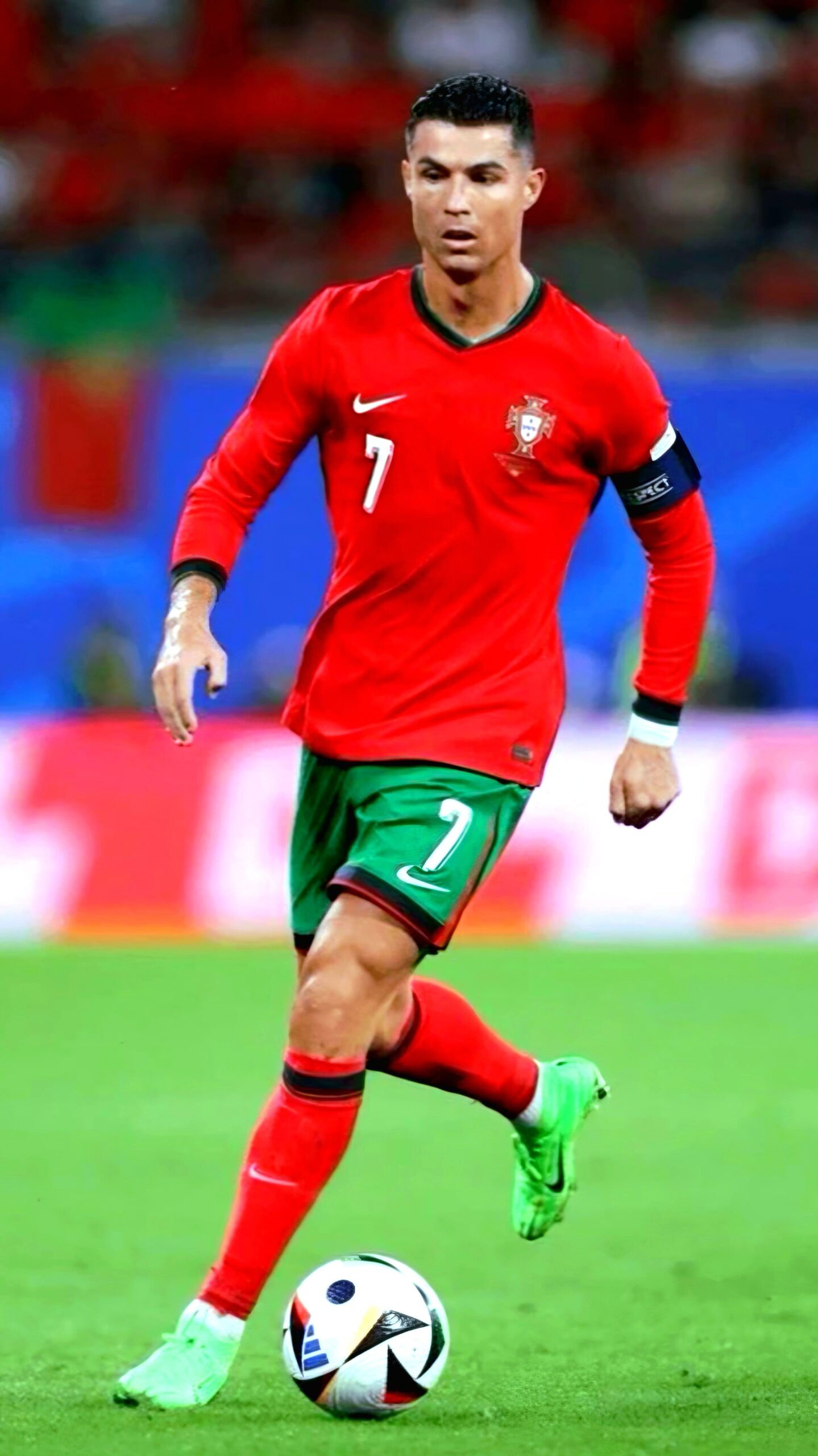 A male soccer player in a red Portugal jersey with number 7 is running on a soccer field, focused on the game, with a ball at his feet. The background is blurred with hints of a crowd, a flag, and IPTV Latvia banners adding to the vibrant atmosphere.