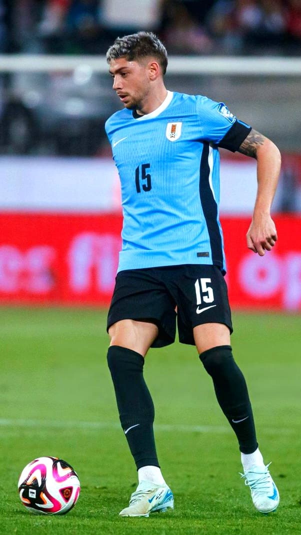 A soccer player in a light blue jersey with number 15 is on the field, focused on controlling a ball with pink, white, and black patterns. He wears black shorts and socks, while the stadium crowd is blurred in the background—a vibrant scene often streamed live via IPTV Latvia for fans who can’t attend in person.